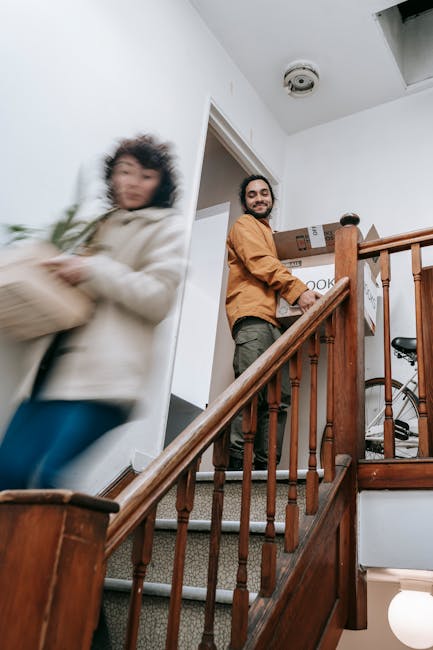 Inside a residential property, a man wearing a brown jacket is standing on a staircase landing holding a cardboard box, preparing for furniture transport or home relocation. The staircase has wooden banisters and carpeted steps, leading to an upper floor. A woman dressed in a white coat and blue jeans is captured in motion walking past the scene, slightly blurred. Nearby, visible in the background, are additional cardboard boxes, some wrapped in plastic or packing paper, indicating packing and moving preparations. The room has white walls, a ceiling with a smoke detector, and natural lighting. The scene illustrates an organized moving process, with furniture and boxes arranged for loading into a removal van, supported by the professional services of Man with Van Gants Hill, specializing in removals and house moves, particularly in urban settings like Gants Hill.