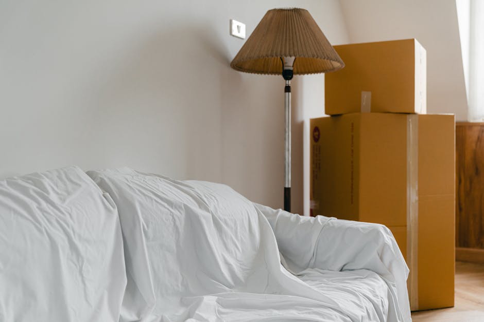 Inside a home room during a house move, three large cardboard moving boxes are stacked next to a wooden cabinet near a window with natural light. In the foreground, a white fabric cover or sheet is dr