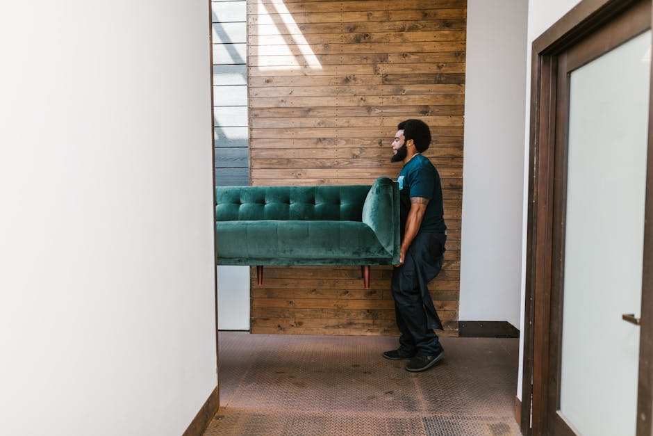A man from Man with Van Gants Hill is engaged in a home relocation process, carrying a large, green upholstered sofa through a doorway. The scene takes place inside a residential property, with a brown wooden wall on one side and white walls on the other, indicating a modern interior. The man, dressed in a dark blue uniform, is positioned on the right side, holding the sofa securely while walking across a brown carpeted floor. Visible inside the room is part of a white piece of furniture or appliance near the sofa. The sofa is partially wrapped in plastic, suggesting it has been packed for transport. Natural light filters into the space, possibly from a window or door area not fully visible in the image. This loading and furniture transport action exemplifies typical steps involved in house removals, with careful handling of bulky items before loading onto a vehicle for a move, supported by [COMPANY_NAME] as part of their removals services.