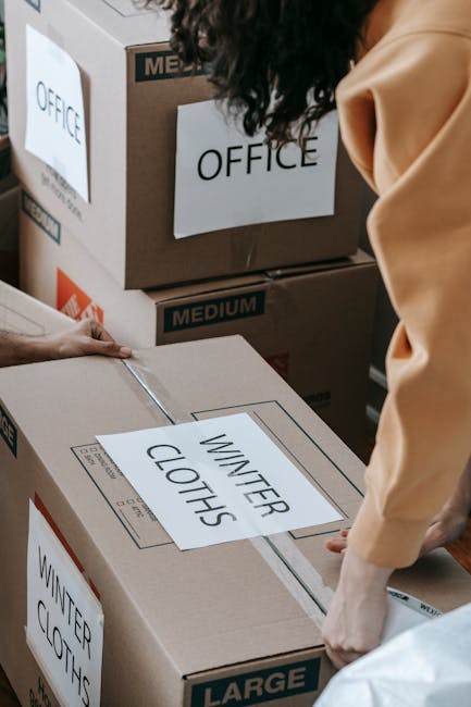 A person wearing a beige jacket is seen handling a large cardboard box labeled 'WINTER CLOTHES' during a home relocation process. The box is positioned on a surface, with other boxes around it, some m