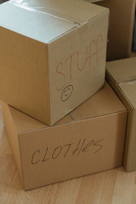 A person wearing a beige jacket is seen handling a large cardboard box labeled 'WINTER CLOTHES' during a home relocation process. The box is positioned on a surface, with other boxes around it, some m