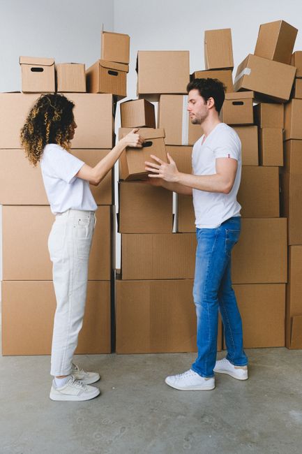 Two individuals are engaged in a packing and loading process within a warehouse or storage area, surrounded by numerous cardboard boxes of various sizes stacked against a plain grey wall. The woman, with curly hair, is dressed in a white t-shirt and light-colored trousers, and is handing or receiving a small cardboard box from the man, who has dark hair and is wearing a white t-shirt and blue jeans. Both are standing on a concrete floor, and the man is holding a slightly larger cardboard box, ready to be loaded into a van or storage space. The environment appears to be an indoor setting with neutral lighting, indicative of a professional home relocation or furniture transport operation. This scene visually supports the logistics involved in house removals, emphasizing packing, handling, and the transportation elements managed by [COMPANY_NAME] during moving or storage services related to house relocations, as highlighted on the page titled 'Confused by Storage Costs in Gants Hill?'