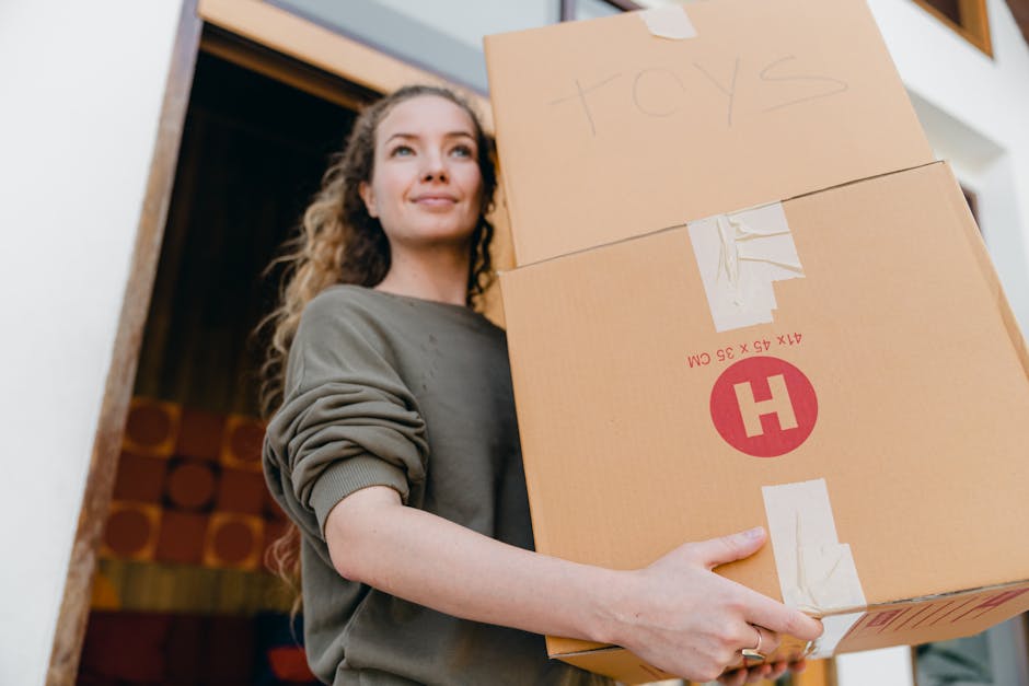 A young woman with curly hair, wearing a grey long-sleeve top, is holding two cardboard boxes during a house relocation. One box, labeled 'toys,' is smaller and positioned on top of a larger box. The larger box features protective packing tape and a red H logo, indicating careful handling. She stands on a paved driveway or porch, with the open doorway of a house visible behind her, revealing wooden paneling inside. There is natural daylight illuminating the scene, highlighting her smiling expression and the condition of the boxes, which are ready for transport by a professional removals service such as Man with Van Gants Hill, specialising in furniture transport and packing during home moves.