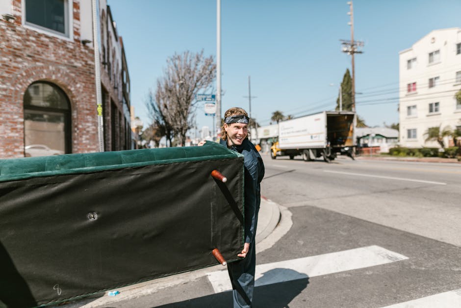 A man wearing a black jacket and a patterned headband is standing outside on a street near a curb, carrying a large, black moving box covered in green fabric with visible wooden handles. Behind him, there is a white van with an open rear door parked on the pavement, suggesting the loading or unloading process during a house removal or furniture transport. The scene is set on a bright day with clear blue skies, and nearby buildings include a brick structure with windows and a white multi-story apartment building in the background. The street features a pedestrian crosswalk, utility poles, and a few trees, indicating an urban environment suitable for professional removals. This image exemplifies a home relocation environment, where furniture and packing materials are being loaded into a van as part of a comprehensive packing and moving operation managed by Man with Van Gants Hill, a company specializing in removals and transport services.
