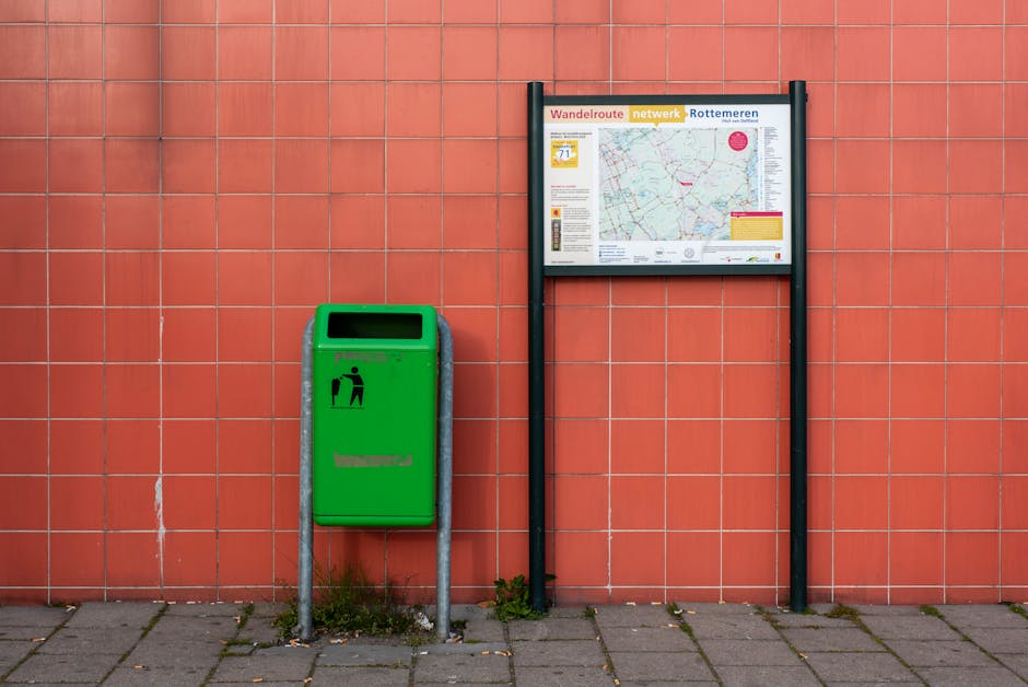 A green public waste bin with a rectangular opening at the top, mounted on a metal frame with four legs attached to a paved sidewalk. The bin features a black icon of a person disposing of trash. To the right of the bin, there is a large outdoor information board displaying a detailed map and various text, supported by two black vertical posts. Both objects are positioned against a red brick wall with a grid-like pattern, indicative of a public space or urban environment. The ground is paved with rectangular grey tiles, and small patches of grass and weeds are visible at the base of the bin. The lighting appears natural, consistent with daytime conditions. This setting is typical of outdoor areas in residential or commercial districts, often associated with public services such as waste disposal or local information, relevant to house removals, packing, and moving logistics support provided by Man with Van Gants Hill.