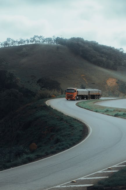 A large orange and black articulated lorry is driving along a winding, two-lane country road surrounded by green grassy hills and sparse trees in the background. The road features sharp curves and a slight incline, with a paved surface marked by white lane lines. The scene appears to be during daytime with natural light, and the landscape suggests a rural area suitable for transport and logistics services, such as those provided by Man with Van Gants Hill, supporting home relocation and furniture transport. The truck is moving away from the camera, positioned on the right side of the road, indicative of UK driving orientation. The surrounding environment is open and peaceful, emphasizing the importance of efficient route planning in house removals and moving logistics.