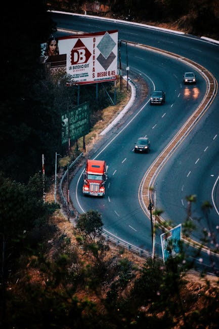 An aerial view of a winding main road with multiple vehicles, including a prominent red truck in the foreground, navigating the curve during daytime. The road features multiple lanes separated by yellow lines, with some vehicles moving in opposite directions. Adjacent to the road, there are green trees and shrubbery, and a large advertising billboard displaying furniture and home-related graphics is situated on the side near the curve. The scene depicts an outdoor setting with natural foliage and clear weather, reflecting typical busy traffic conditions that could be encountered during home relocation or furniture transport logistics. The image supports content related to efficient routes for house removals, as showcased on the webpage for Man with Van Gants Hill, which specializes in house removals and moving services in Ilford and the surrounding areas.