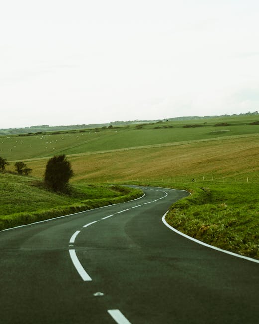 A large orange and black articulated lorry is driving along a winding, two-lane country road surrounded by green grassy hills and sparse trees in the background. The road features sharp curves and a slight incline, with a paved surface marked by white lane lines. The scene appears to be during daytime with natural light, and the landscape suggests a rural area suitable for transport and logistics services, such as those provided by Man with Van Gants Hill, supporting home relocation and furniture transport. The truck is moving away from the camera, positioned on the right side of the road, indicative of UK driving orientation. The surrounding environment is open and peaceful, emphasizing the importance of efficient route planning in house removals and moving logistics.