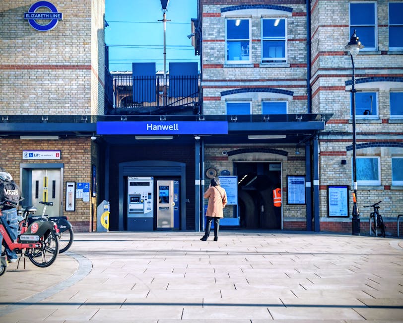 Exterior view of Hanwell underground station entrance in daylight, with a woman standing on the pavement facing the ticket machines and entrance archway. The station facade is constructed with light-colored bricks and features a dark blue sign displaying 'Hanwell' above the entrance, along with official signage for the Elizabeth Line at the top left corner. To the left of the entrance, there are bike-sharing bikes from Uber and VCS, secured to bike racks. Several information and ticket vending machines are positioned directly in front of the station entrance, with a small covered area covering the doorway and an illuminated security camera on the right side. The pavement in front is clean and paved, and a street lamp with a classic lantern-style light fixture is mounted on the building's corner. The scene suggests a typical urban setting, potentially used for home relocation planning or moving logistics involving transport to or from the station, with [COMPANY_NAME] providing removals services in the vicinity.