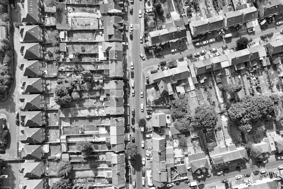 An aerial black-and-white photograph showing a narrow urban street surrounded by terraced houses with small backyards, some with visible garden furniture, trees, and fences. The street is lined with parked cars on both sides, and a few vehicles or vans are present, some close to the curbs. Additionally, a manwithvangantshill.co.uk removal van is parked on the pavement near the house entrances, with a few cardboard boxes and packing materials visible on the sidewalk. The scene captures a house removal or packing and moving process as part of a home relocation, with the loading process ongoing. The environment is well-lit, with shadows indicating a sunny day, and the overall setting reflects a typical residential neighbourhood in Gants Hill, suitable for professional removals services by Man with Van Gants Hill.