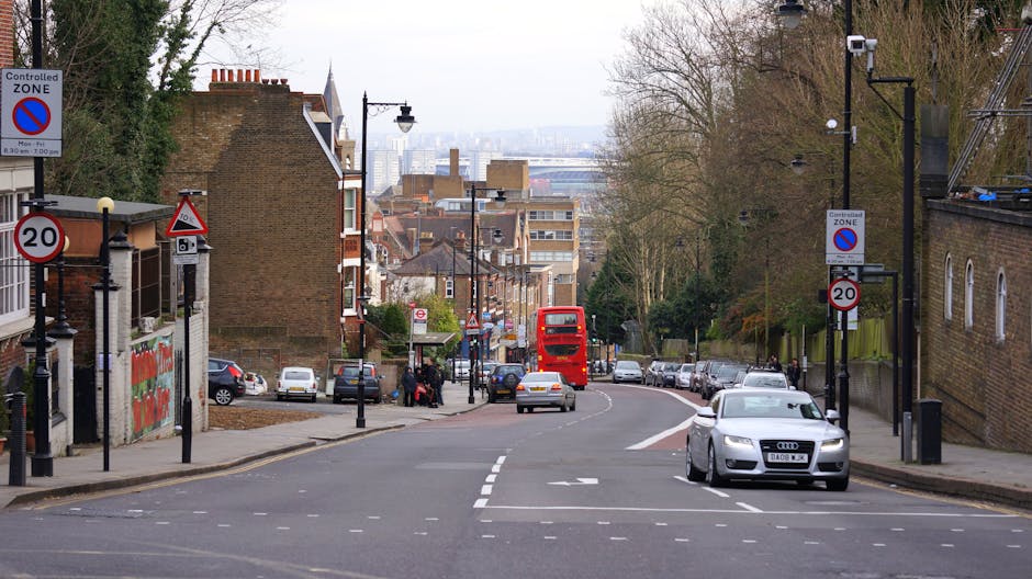 A street scene in Gants Hill with a designated controlled zone sign visible on the sidewalk, indicating parking restrictions during certain hours. Several parked cars line both sides of the road, including a white vehicle in the foreground and other vehicles further down the street. A red double-decker bus is approaching, traveling away from the viewer. The street curves gently to the right, with a bus stop shelter and several pedestrians standing near it on the pavement. Residential buildings and brick houses are visible on the left, while on the right side, there are trees, lampposts, and a brick wall. Overhead streetlights and power lines are also present, with a glimpse of the city skyline in the distance under an overcast sky. This scene depicts typical urban movement and parking conditions, which are relevant to planning house removals or moving services in the area, as seen on the page titled 'Redbridge Council Permits for Gants Hill Street Moves' with efforts by Man with Van Gants Hill in facilitating efficient furniture transport and home relocation processes.