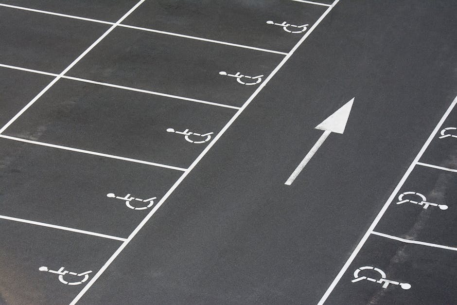 A section of an outdoor parking lot with clearly marked designated parking spaces, each outlined with white paint and featuring wheelchair-accessible symbols. The parking spaces are arranged along the curb, with an arrow painted on the asphalt indicating the direction of traffic flow moving forward through the lot. The surface of the lot appears smooth and clean, with no vehicles visible. The scene suggests a quiet, organized area suitable for household or business relocations, where careful planning for vehicle parking and access is essential. In the context of home relocation and furniture transport, Man with Van Gants Hill can assist with planning parking arrangements and ensuring smooth loadings and unloadings during moving day, taking accessibility and parking regulations into account.