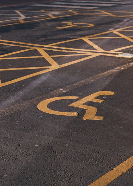 A section of an outdoor parking lot with clearly marked designated parking spaces, each outlined with white paint and featuring wheelchair-accessible symbols. The parking spaces are arranged along the curb, with an arrow painted on the asphalt indicating the direction of traffic flow moving forward through the lot. The surface of the lot appears smooth and clean, with no vehicles visible. The scene suggests a quiet, organized area suitable for household or business relocations, where careful planning for vehicle parking and access is essential. In the context of home relocation and furniture transport, Man with Van Gants Hill can assist with planning parking arrangements and ensuring smooth loadings and unloadings during moving day, taking accessibility and parking regulations into account.